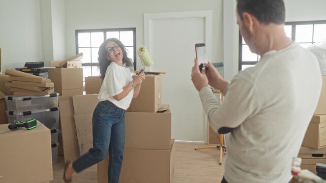 Man photographing smiling woman holding paint roller and showing peace sign with smartphone among moving boxes inside building; playful teamwork.