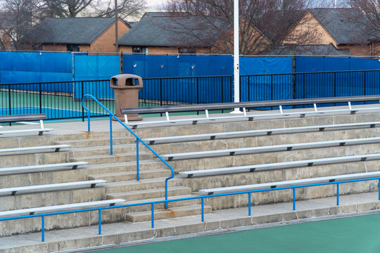 Empty stadium bleachers and tennis court with blue fence, trash can, and railings, awaiting spectators for a match or event, showcasing a quiet sports facility.