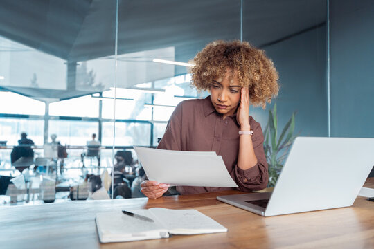 Overwhelmed stressed office worker woman worried reading business documents at desk with laptop in modern workplace, hand on head, paperwork and notebook on table, corporate setting. Copy space