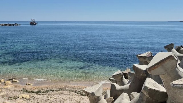 ​A scenic coastal view featuring a sandy beach, clear turquoise water, and a stack of large concrete tetrapods in the foreground. A pirate-style ship is near a rocky outcrop in the distance.