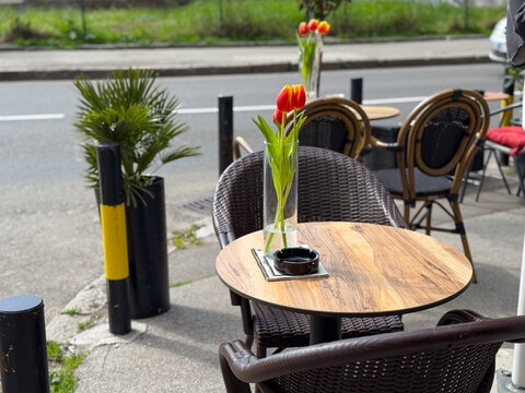 Cafe table with glass vase holding tulips on a sidewalk terrace near street. Outdoor dining setup, hospitality service, relaxation moment and small business atmosphere.