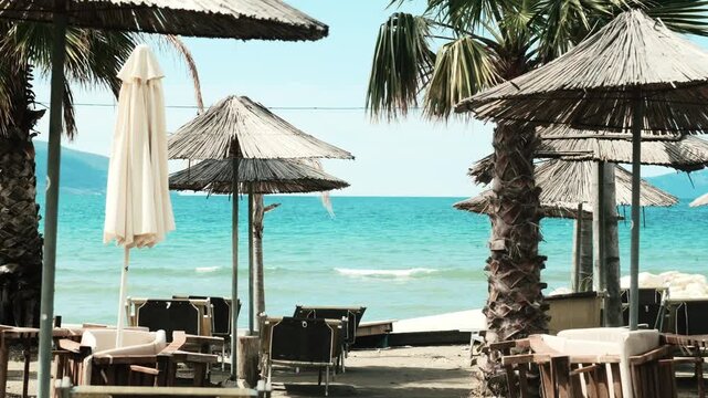 Handheld Tropical beach with straw parasols, palm trees and empty lounge chairs near turquoise sea. Sunny seaside resort scene with wooden furniture and closed umbrellas, calm water and summer travel 
