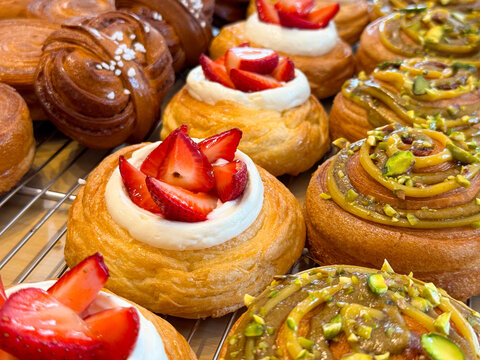 Danish pastries with strawberry cream and pistachio topping arranged closely on bakery rack. Sweet dessert selection, pastry variety, confectionery display and food styling concept.