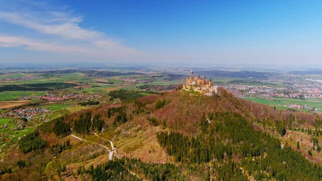 Burg Hohenzollern, Germany - 15 April 2026: Aerial view of Hohenzollern Castle perched on a hilltop surrounded by lush forests and rolling green fields under a clear blue sky.