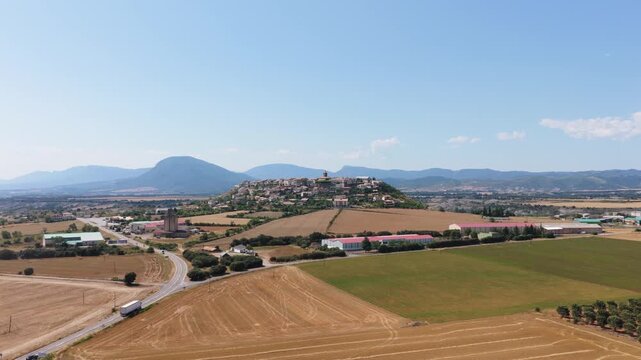 Aerial view tracking a large truck driving on a country road with the historic hilltop town of Berdun in the background on a sunny day