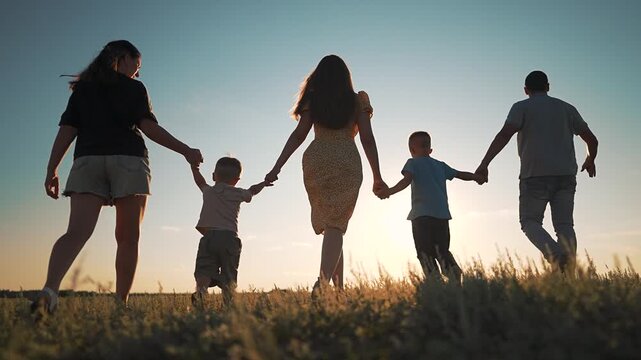 Happy family is walking in green field at sunset. Parents and children hold hands in a row. Group of people enjoys summer evening in nature. Family walks away together in nature park happily.