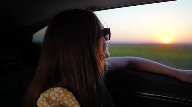 Young woman in sunglasses travels by car at sunset. Happy traveler looks out car window during summer road trip. Girl enjoys sunset travel in passenger seat. Vacation car ride. She smiles.