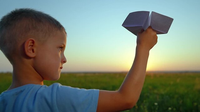 Boy holds paper airplane overhead in field at sunset. Child plays with paper plane outdoors. Kid preparing to fly aircraft. Boy with paper glider at golden hour. Child plays with airplane outside.