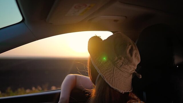 Young woman passenger looks out car window at sunset. Girl traveler enjoys summer road trip in vehicle. Female in hat watches golden sun during car ride. Happy woman goes on vacation journey.