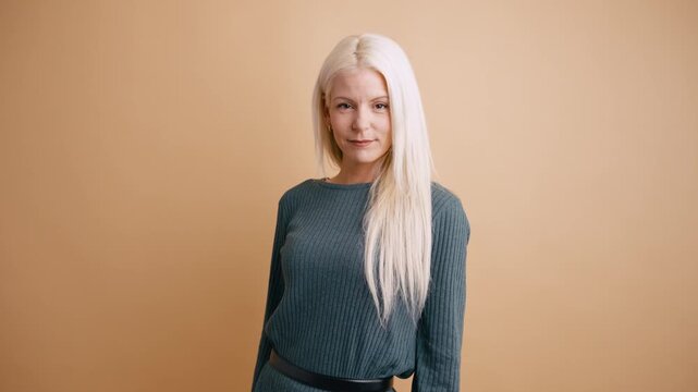 Beautiful mature woman with long straight platinum blonde hair looking at the camera and smiling gently, posing in a studio with a beige background