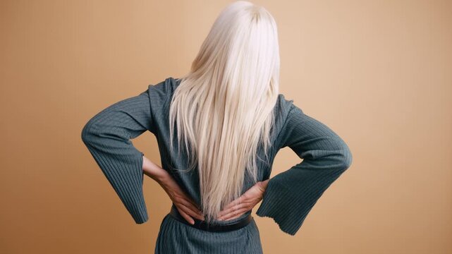 Unrecognizable blonde woman suffering from intense back pain. She is standing with her back to the camera, massaging her lower back