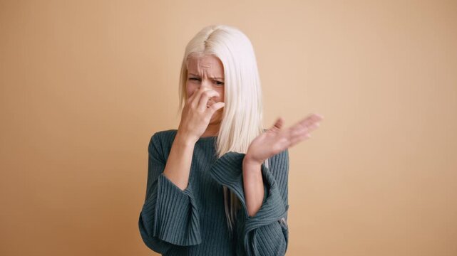 Young blonde woman smelling something stinky and disgusting. She pinches her nose with her fingers and waves her hand, expressing disgust