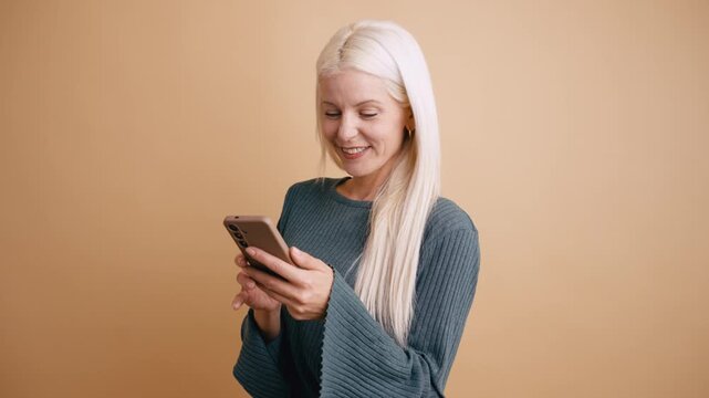 Smiling blonde woman happily using her smartphone. Middle aged lady looking at her phone, scrolling and typing against a beige background