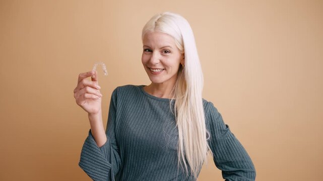 Beautiful middle aged woman with blonde hair holding a transparent orthodontic retainer. Smiling patient showing her removable braces