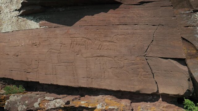 Static handheld shot showing ancient animal petroglyphs carved into a reddish rock surface. Sunlight illuminates the weathered, cracked stone, revealing the prehistoric drawings.