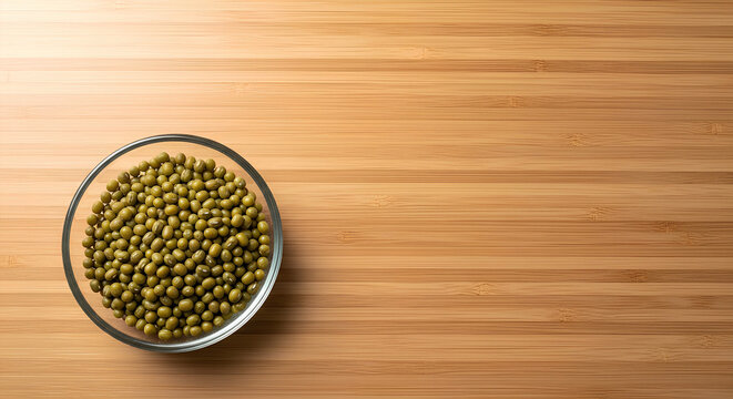 Small glass dish filled with dry mung beans placed in the lower left corner of a large light bamboo surface, most of the bamboo kept empty as negative space for Asian pantry or sprouting recipe text.