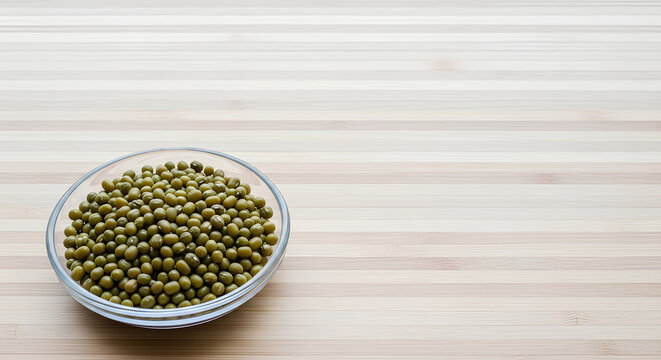 Small glass dish filled with dry mung beans placed in the lower left corner of a large light bamboo surface, most of the bamboo kept empty as negative space for Asian pantry or sprouting recipe text.