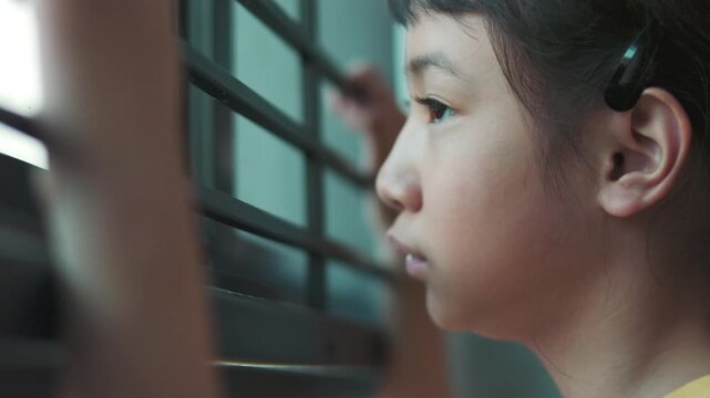 Close up side profile of sad Asian girl looking out window with selective focus. Lonely child face details with blurred hand holding bars in foreground, concept of waiting, depression, or boredom.