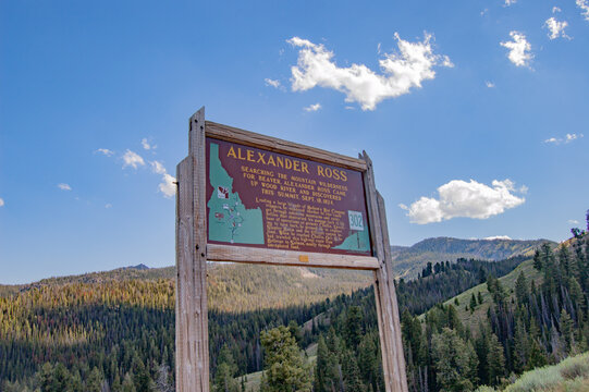 Wooden sign marks Alexander Ross trailhead. Sky shows blue with scattered white clouds. Green mountains surround the sign. Trees cover the rolling hills. Sign features map and trail information