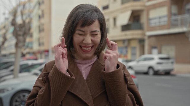 Woman smiling with fingers crossed and eyes closed on an urban street amid parked cars and apartment buildings; hope luck.