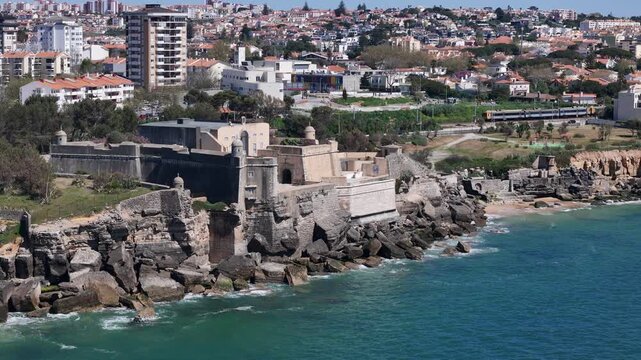 Estoril, Portugal - 30 March 2026: Aerial view of Forte de Santo Antonio da Barra on the rocky coastline with the town and a train in the background.