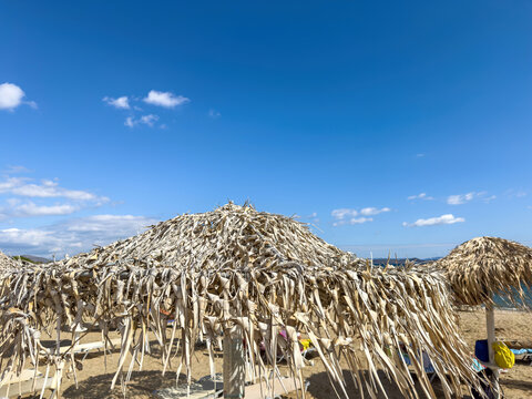 Straw beach umbrella on sandy shore by calm Aegean Sea, Attica, Greece. Bright sun and blue sky, relaxation by the sea