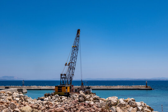 Floating crane barge working near rocky pier in turquoise Greek island harbor under blue summer sky