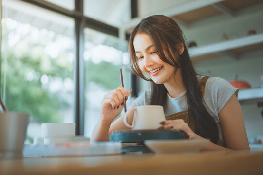Happy young Asian woman painting a white ceramic mug with a brush in a pottery studio for creative hobby and handmade craft concept