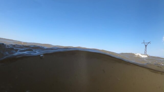 A unique half underwater view captures small rolling waves in a murky lake. A piece of driftwood sticks out while mountains and trees line the distant sandy shore.

