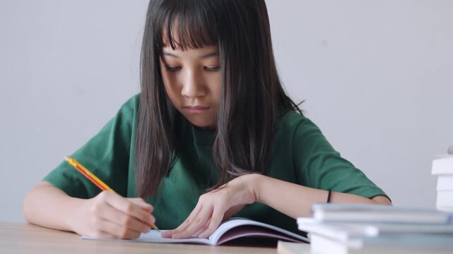 Close up selective focus of a concentrated Asian kid, a young Thai girl or Chinese student, diligently doing homework and writing with a pencil in a notebook. Education, learning, and study concept.