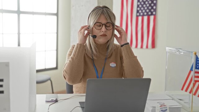 Woman wearing headset adjusts glasses at laptop beside a clear ballot box with voted sticker in a polling station building; frustration.
