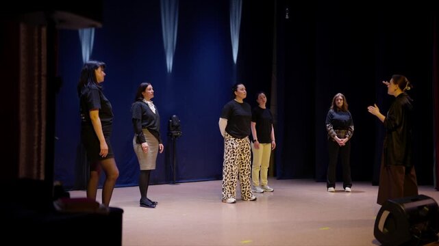 Theater instructor guiding diverse group of young performers on stage during rehearsal with dramatic lighting, focusing on communication, staging, and ensemble presence