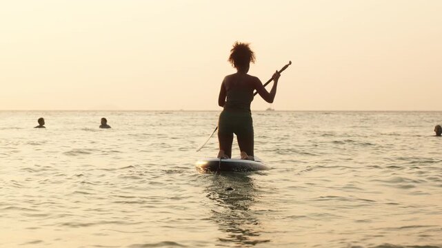 Silhouette of a woman paddleboarding on calm sea during golden sunset, reflecting sunlight on water with distant boat on horizon.