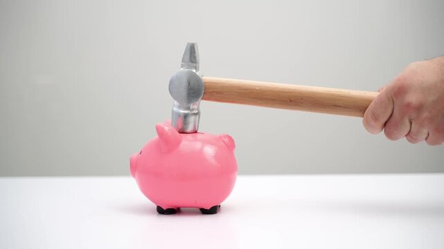Pink piggy bank sits on white surface. Hammer hovers above it, poised to strike. Hand grips the hammer's wooden handle. No facial expression visible, but intent is clear