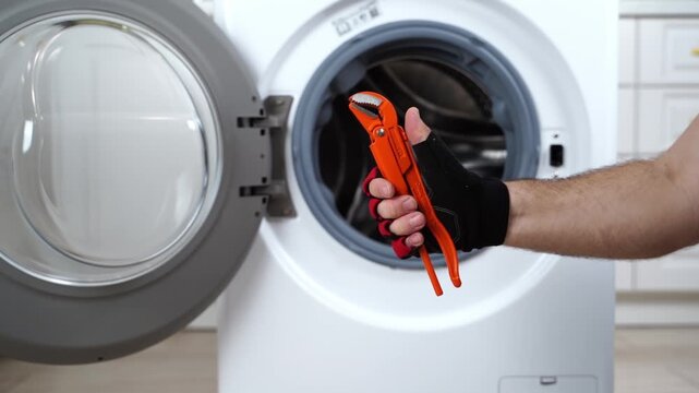 Open door reveals empty washing machine drum. Hand enters frame holding red-handled pliers. Person grips tools, ready for repair work. Scene implies maintenance or fixing of home appliance