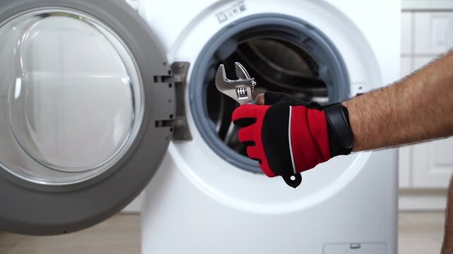 Hand holds wrench near washing machine door. Open door shows inner drum. Wrench suggests repair or maintenance. Calm action implies practical problem-solving. Use for home repair or appliance content