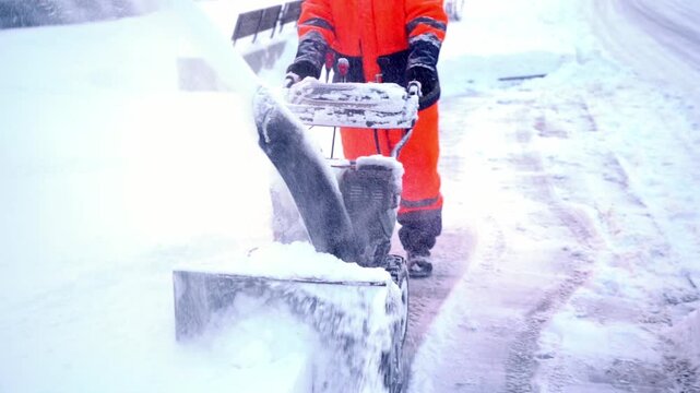 Snowplow operator clears snow from a sidewalk. Bright orange gear contrasts white snow. Snow blasts upward as blade moves forward. Person focused on clearing path quickly