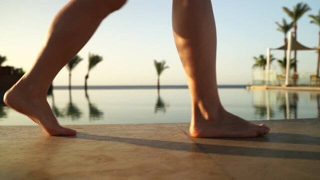 Feet step barefoot near infinity pool. Sunlight glows warm and golden. Reflections ripple gently on water. Palm trees sway softly in breeze. Calm scene evokes relaxation and peace