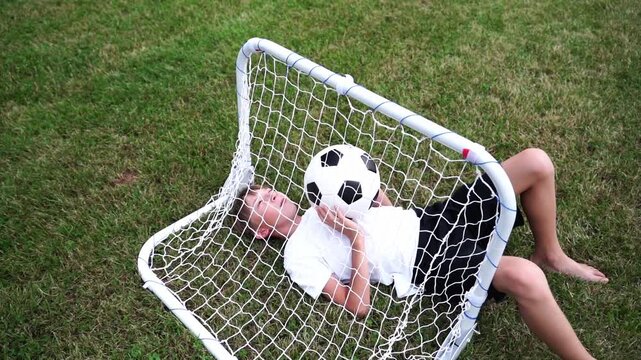 Child rests in soccer goal, holding ball. Child smiles, lying on grass, eyes closed. Child's playful pose shows joyful relaxation. Perfect for sports, parenting, or child activity themes