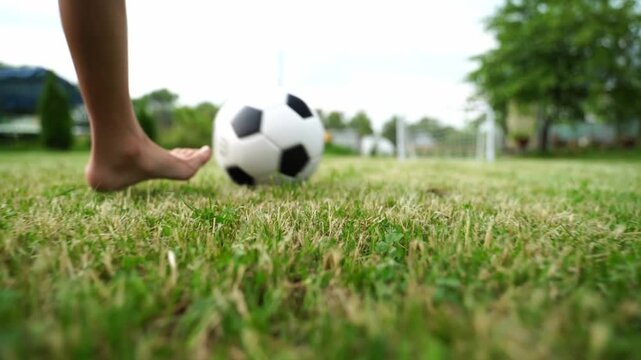 Child's bare foot near soccer ball on green grass. Soft focus shows trees and distant goal. Natural daylight enhances outdoor play feel. Great for youth sports or active lifestyle themes
