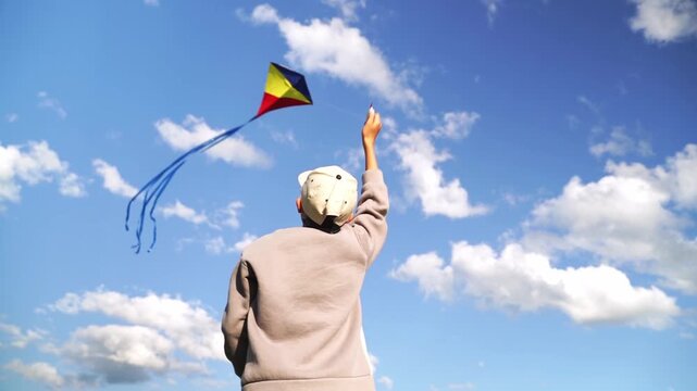 Child flies kite against bright blue sky. Wearing helmet, child smiles, arms raised high. Kite dances above, tails fluttering in wind. Perfect for outdoor, joyful, nostalgic, or childhood themes