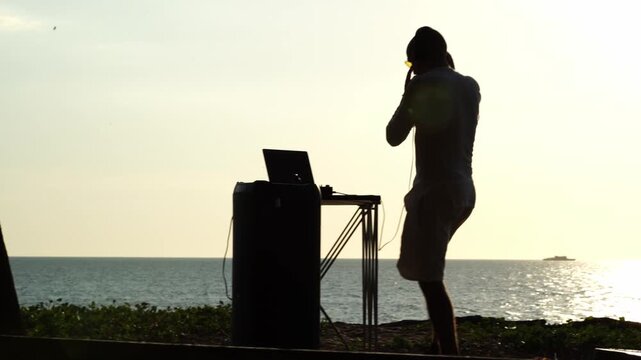 Silhouetted DJ mixes music by sea at golden hour. He gestures with headphones, eyes focused on screens. Ocean waves crash softly behind his portable setup