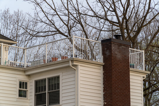 Residential house featuring a spacious rooftop deck with white railings, a prominent red brick chimney, white siding, and windows, set against bare tree branches in early spring, showcasing home archi