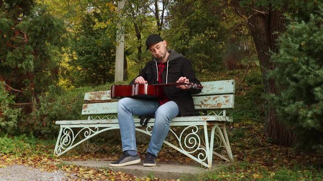 Wide shot of young Caucasian man is sitting on a park bench and unusually playing the guitar