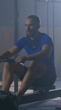 Focused male athlete training on indoor rowing machine during intense workout session in dimly lit gym, transitioning from rest to powerful pulling motion