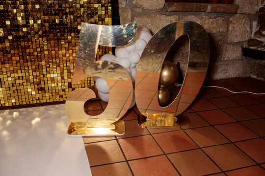 Large shiny gold mirror number 50 figures filled with white and chrome gold balloons stand on a tile floor against a glittering sequin wall at a birthday party