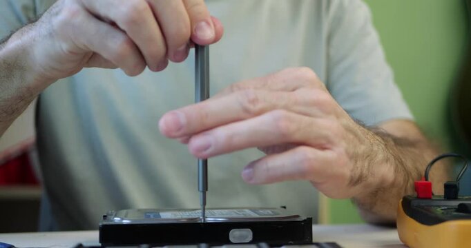 Technician repairing hard drive with screwdriver in workshop, close-up of hands working on device. Small business IT service, data recovery and hardware maintenance, professional electronics repair.
