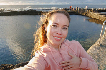Fototapeta premium A woman smiles as she takes a selfie at sunset by the sea