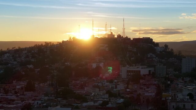 Stunning sunset illuminates Durango Mexico as golden light cascades over densely packed hillside buildings. Radio transmission towers silhouette against warm orange sky with lens flare.