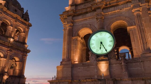 Cathedral Basilica of Immaculate Conception in Durango, Mexico illuminated at dusk with ornate baroque towers and glowing clock face against deep blue evening sky with cityscape.